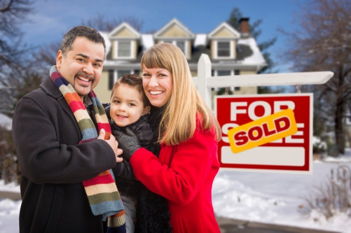 Warmly Dressed Young Mixed Race Family in Front of Sold Home For Sale Real Estate Sign and House with Snow On The Ground.