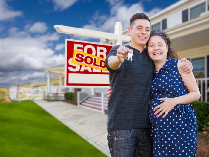 Happy Hispanic Couple In Front of New Home And Sold Real Estate Sign Showing Off Their House Keys.